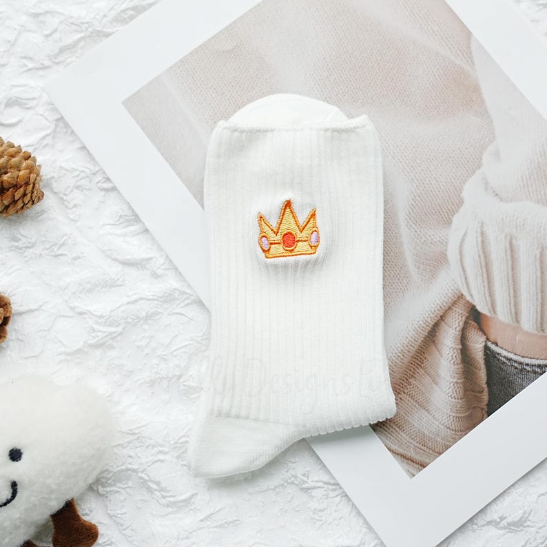 May include: White ribbed socks with an embroidered gold and orange crown design. The socks are made of a soft material and are folded. The crown is centered on the sock. The socks are a light cream color.