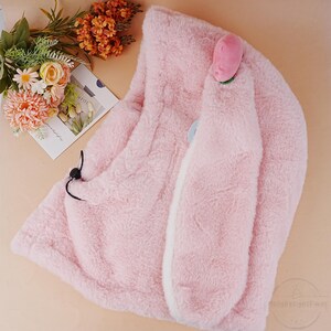 May include: A pink plush hooded scarf with bunny ears and a pink flower on top. The scarf is made of soft, fuzzy material and has a white lining.