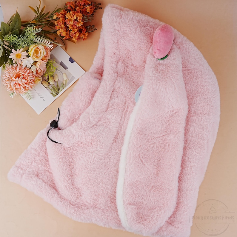 May include: A pink plush hooded scarf with bunny ears and a pink flower on top. The scarf is made of soft, fuzzy material and has a white lining.
