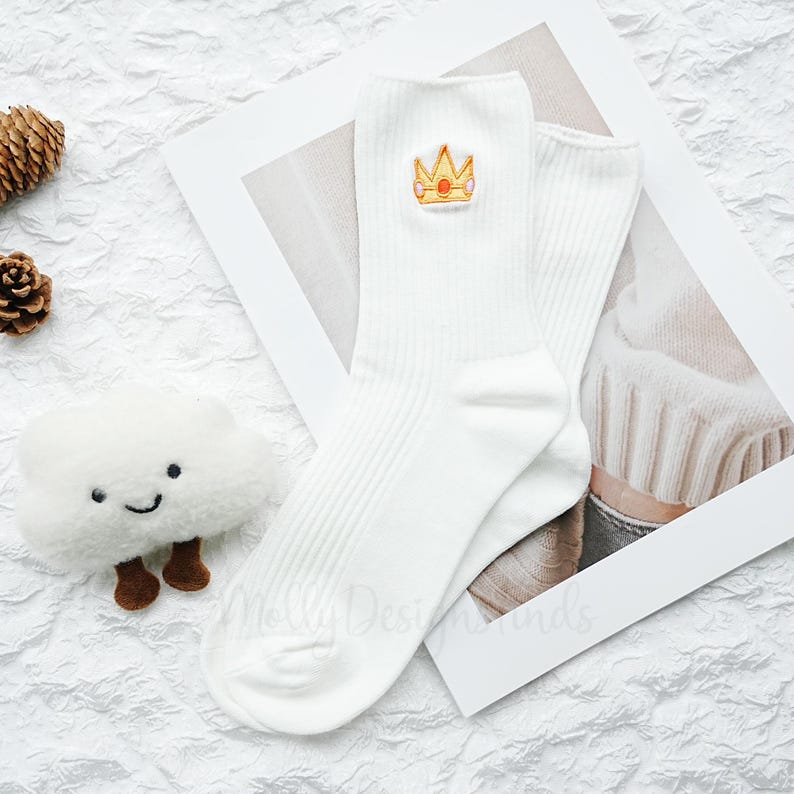 May include: A pair of white ribbed socks with a gold and red crown patch. The socks are folded on a white background with a small plush cloud and pinecones.