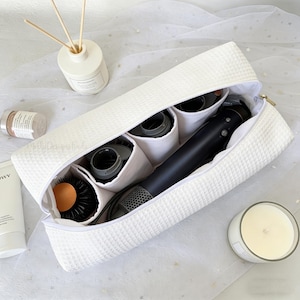 May include: A white, textured travel case with a gold zipper, open to reveal a black hair dryer and attachments. The case has internal compartments for organization. A round brush and other accessories are visible.