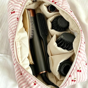 Könnte beinhalten: Eine rosa-weiß gestreifte, gesteppte Kosmetiktasche mit Kirschmuster, geöffnet, um verschiedene Haarstyling-Tools zu enthüllen. Die Tasche enthält ein Glätteisen, Bürsten und anderes Zubehör, alles ordentlich organisiert.