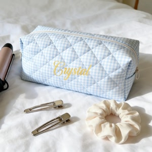 May include: A light blue and white gingham quilted cosmetic bag with the name "Crystal" embroidered in yellow. Two silver barrettes and a cream-colored scrunchie are also visible.