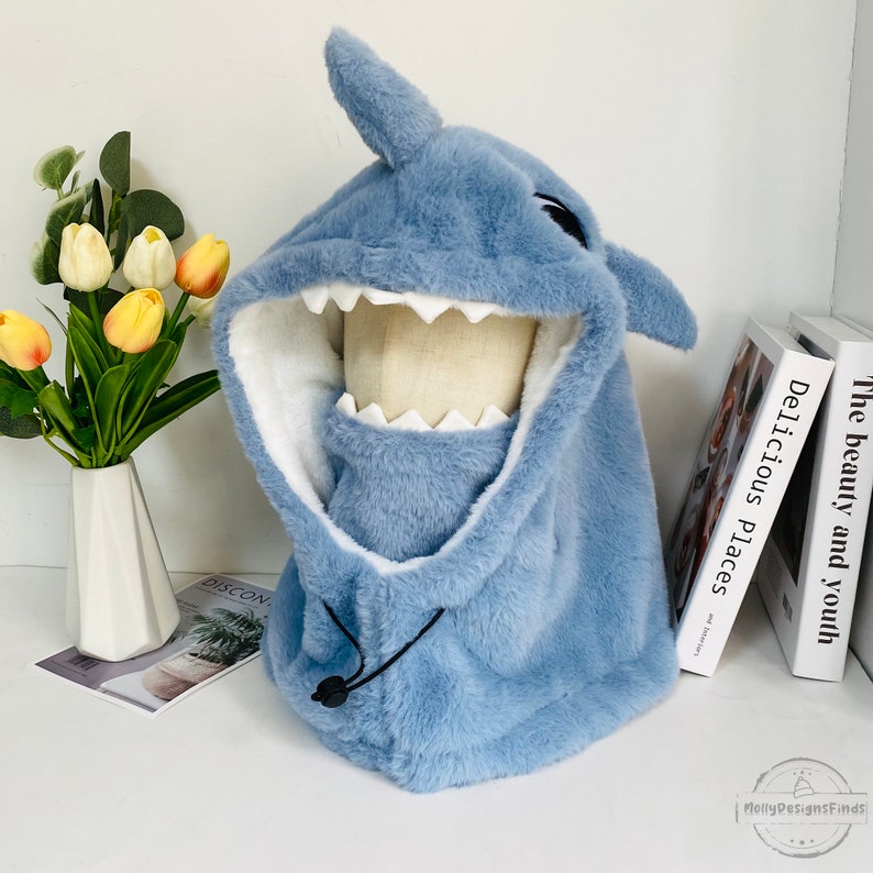 May include: A light blue plush shark hooded hat with a white lining and a black drawstring. The hat has a large, toothy grin and a pair of black eyes.