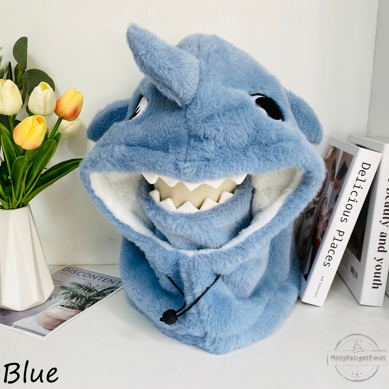 May include: Blue plush shark hooded neck warmer with a white mouth and teeth. The neck warmer has a drawstring closure.