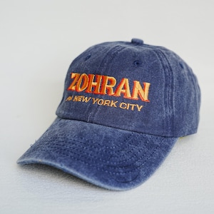 Pu&ograve; includere: Un berretto da baseball in denim blu con la scritta "ZOHRAN" ricamata in lettere arancioni. Sotto la parola "ZOHRAN" c'&egrave; il testo "NEW YORK CITY" in lettere arancioni. Il berretto ha una visiera curva e un bottone sulla parte superiore.