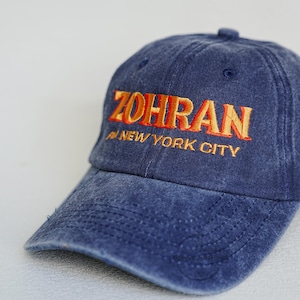 Pu&ograve; includere: Cappellino da baseball in denim blu con la scritta "ZOHRAN" ricamata in lettere arancioni. Sotto, il testo "NEW YORK CITY" &egrave; anch'esso arancione. Il cappellino ha una visiera curva e un bottone sulla parte superiore.