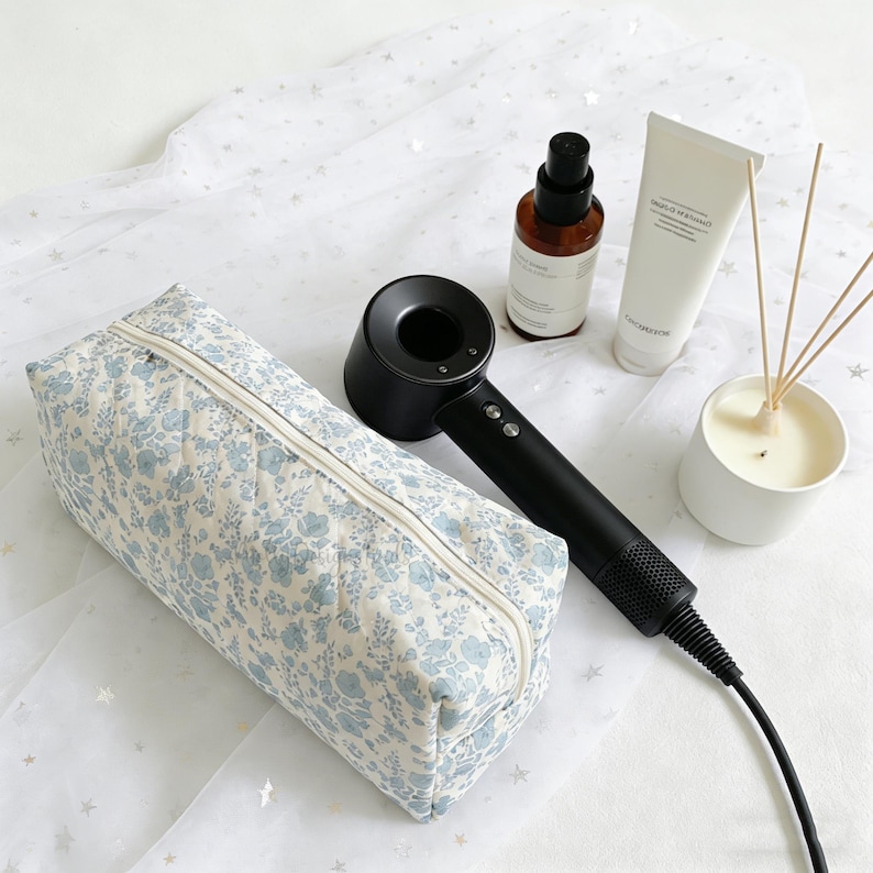 May include: A floral print cosmetic bag with a zipper, a black hair dryer, a brown bottle, a white tube, and a candle with reeds. The cosmetic bag has a light blue floral pattern on a cream background. The hair dryer is black.