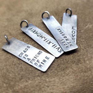 May include: Five silver metal tags with different inscriptions. The tags are rectangular and have rounded corners. The inscriptions are: "Chaos is a friend of mine", "XVII: MCMXIV", "MEMORIAS MORRIAS", and "LOVE ME NOW".