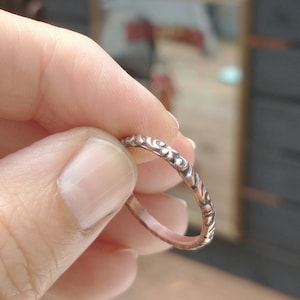 May include: A thin copper ring with a detailed floral pattern. The ring is shown on a finger.