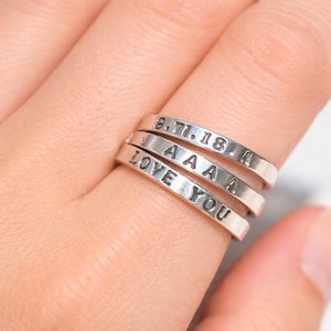 May include: Silver rings stacked on a finger, each engraved with text. The top ring displays "9.7.18.4", the second "A A A A", and the third "LOVE YOU". The rings are a simple, elegant design.