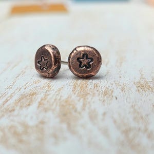 May include: A pair of copper-colored stud earrings. Each earring has a textured, rounded surface with a star-shaped design. The earrings are displayed on a light-colored, textured surface.