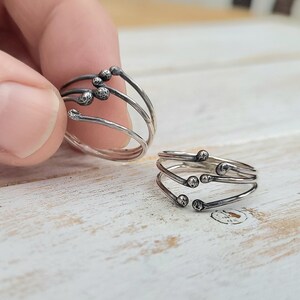 Puede incluir: Dos anillos de plata con un dise&ntilde;o &uacute;nico. Cada anillo presenta m&uacute;ltiples bandas con peque&ntilde;as esferas texturizadas en los extremos. Uno est&aacute; sostenido por una mano, mientras que el otro descansa sobre una superficie de madera clara.