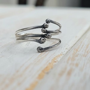 Puede incluir: Un anillo plateado con un dise&ntilde;o &uacute;nico. El anillo presenta una banda enrollada con cinco extremos en forma de cerilla, cada uno con una cabeza texturizada. El anillo se encuentra sobre una superficie de madera blanca, creando un contraste.