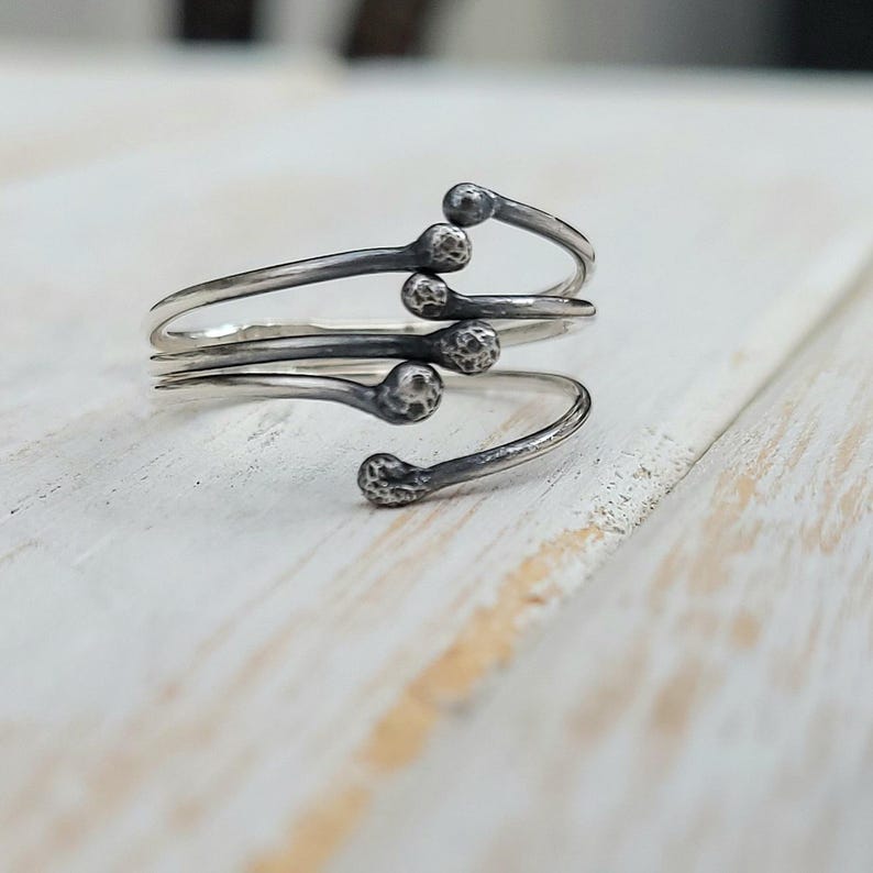 Puede incluir: Un anillo plateado con un dise&ntilde;o &uacute;nico. El anillo presenta una banda enrollada con cinco extremos en forma de cerilla, cada uno con una cabeza texturizada. El anillo se encuentra sobre una superficie de madera blanca, creando un contraste.