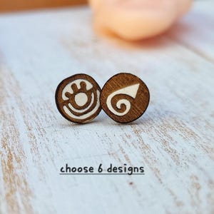 May include: Two circular wooden stud earrings featuring dark brown wood grain with engraved white designs. One earring displays a stylized eye symbol, while the other shows a spiral arrow icon. The text choose 6 designs is visible.