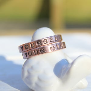 May include: Two copper-coloured metal rings stacked on top of each other. The top ring reads "YOURSELF" and the bottom ring reads "YOUR MIND" in a sans-serif font. The rings are resting on a white ceramic bird figurine.