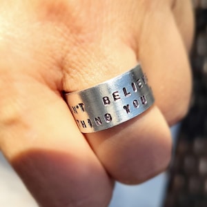 May include: A wide silver-toned metal ring featuring stamped black lettering that reads "DON'T BELIEVE EVERYTHING YOU". The band has a brushed finish and the text is clearly indented into the surface of the metal, creating a high-contrast appearance.