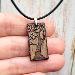 May include: Wooden pendant necklace featuring a rectangular pendant with a carved tree design. The pendant is a light brown color and hangs from a black cord. The tree design is detailed with visible leaves and branches.
