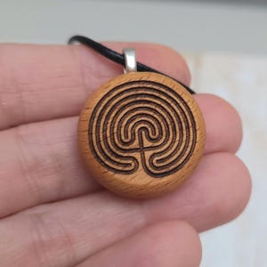 May include: A round wooden pendant with a labyrinth design carved into its surface. The pendant is light brown with dark brown etched lines and hangs from a black cord. A silver-colored metal loop connects the pendant to the cord.