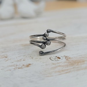 Puede incluir: Un anillo plateado con un dise&ntilde;o &uacute;nico. El anillo presenta una forma en espiral con m&uacute;ltiples brazos, cada uno terminando en un detalle redondeado y texturizado. El anillo se muestra sobre una superficie clara y texturizada.