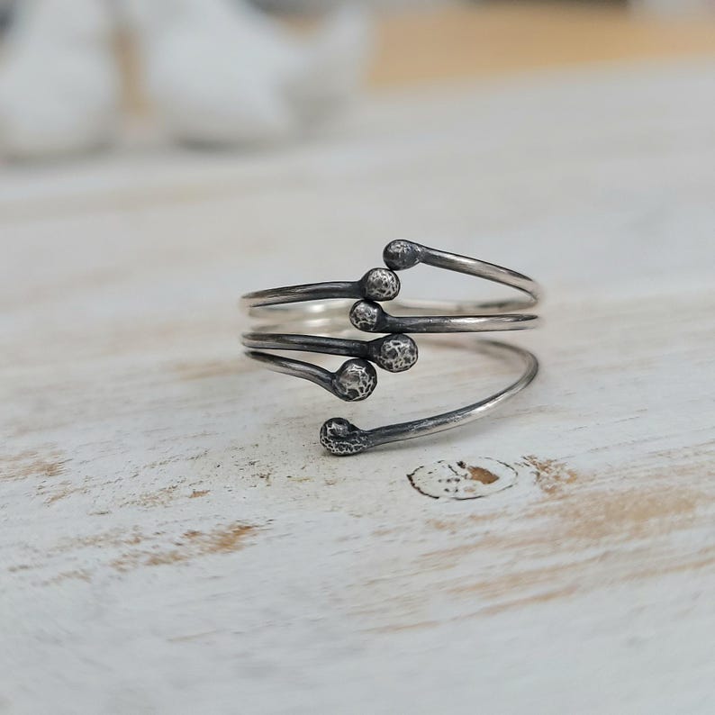 Puede incluir: Un anillo plateado con un dise&ntilde;o &uacute;nico. El anillo presenta una forma en espiral con m&uacute;ltiples brazos, cada uno terminando en un detalle redondeado y texturizado. El anillo se muestra sobre una superficie clara y texturizada.
