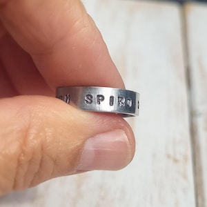 May include: A silver-toned metal band ring featuring the stamped text "SPIRO" in capital letters. The ring has a flat, smooth surface with a metallic finish and clearly visible, indented black lettering across the front of the band.