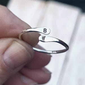 May include: A silver adjustable ring featuring the letters "B" and "W" engraved on the ends. The ring has a minimalist design and is shown against a soft, blurred background.