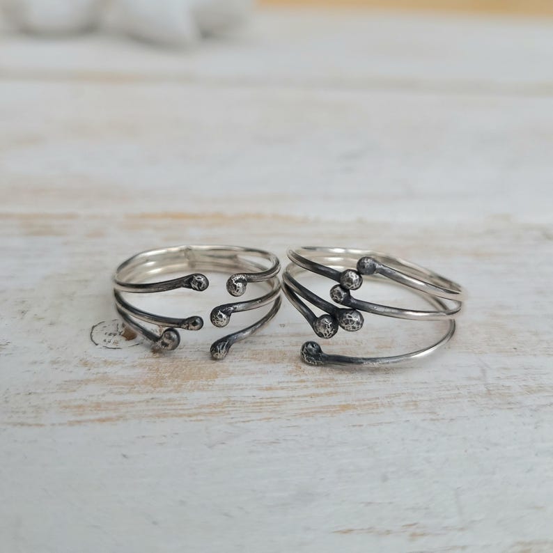 Puede incluir: Dos anillos de plata con un dise&ntilde;o &uacute;nico. Cada anillo presenta m&uacute;ltiples bandas curvas y peque&ntilde;os adornos redondos. Los anillos se exhiben sobre una superficie clara y texturizada, mostrando sus intrincados detalles y artesan&iacute;a.