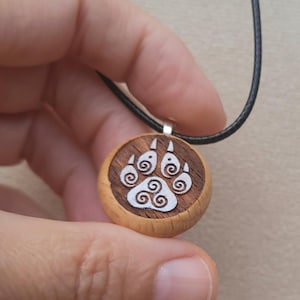 May include: A round wooden pendant with a carved white paw print design. The pendant is attached to a black cord necklace. The pendant is held in a hand.