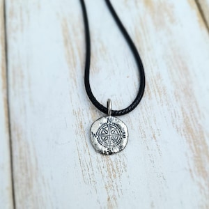 May include: A silver-colored compass pendant necklace with a black cord. The pendant features a detailed compass rose design with directional markings. The necklace is displayed against a light-colored wooden surface.