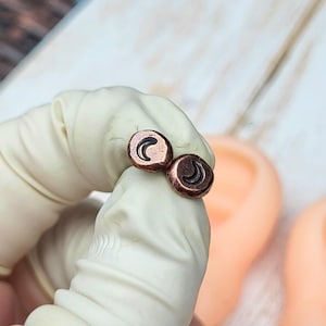 May include: A pair of small, round copper earrings. Each earring features a crescent moon design in black. The earrings are held by a gloved hand, with a blurred background.