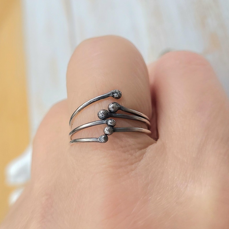 Puede incluir: Un anillo plateado con un dise&ntilde;o &uacute;nico. El anillo presenta cuatro bandas curvas, cada una con una peque&ntilde;a bola texturizada en el extremo, que se asemejan a cerillas. El anillo se lleva en un dedo, mostrando su estilo art&iacute;stico e inusual.