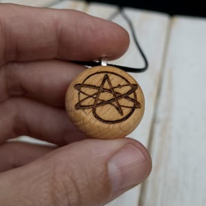 May include: A wooden pendant with a black cord. The pendant is round and made of light-colored wood. It features a dark brown etched design of a star within a circle and intersecting lines.