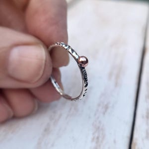 May include: A silver ring with a textured band and a small, copper-coloured ball accent. The ring is held up close, showing the details of the metalwork and the contrast between the silver and copper.
