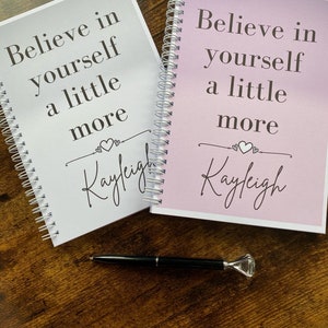 May include: Two white spiral bound notebooks with the text "Believe in yourself a little more" and the name "Kayleigh" written in cursive. The notebooks are on a wooden surface with a black pen.