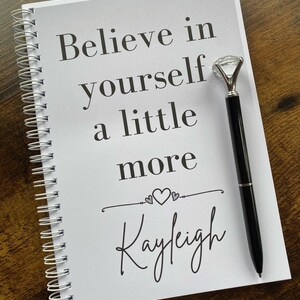 May include: A white spiral bound notebook with the text "Believe in yourself a little more" and the name "Kayleigh" written in cursive. A pen with a diamond-shaped topper is resting on the notebook.