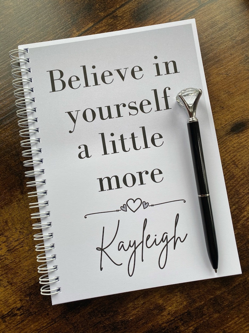 May include: A white spiral bound notebook with the text "Believe in yourself a little more" and the name "Kayleigh" written in cursive. A pen with a diamond-shaped topper is resting on the notebook.