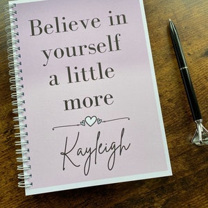 May include: A pink and white spiral bound journal with the text "Believe in yourself a little more" and the name "Kayleigh" written in cursive.