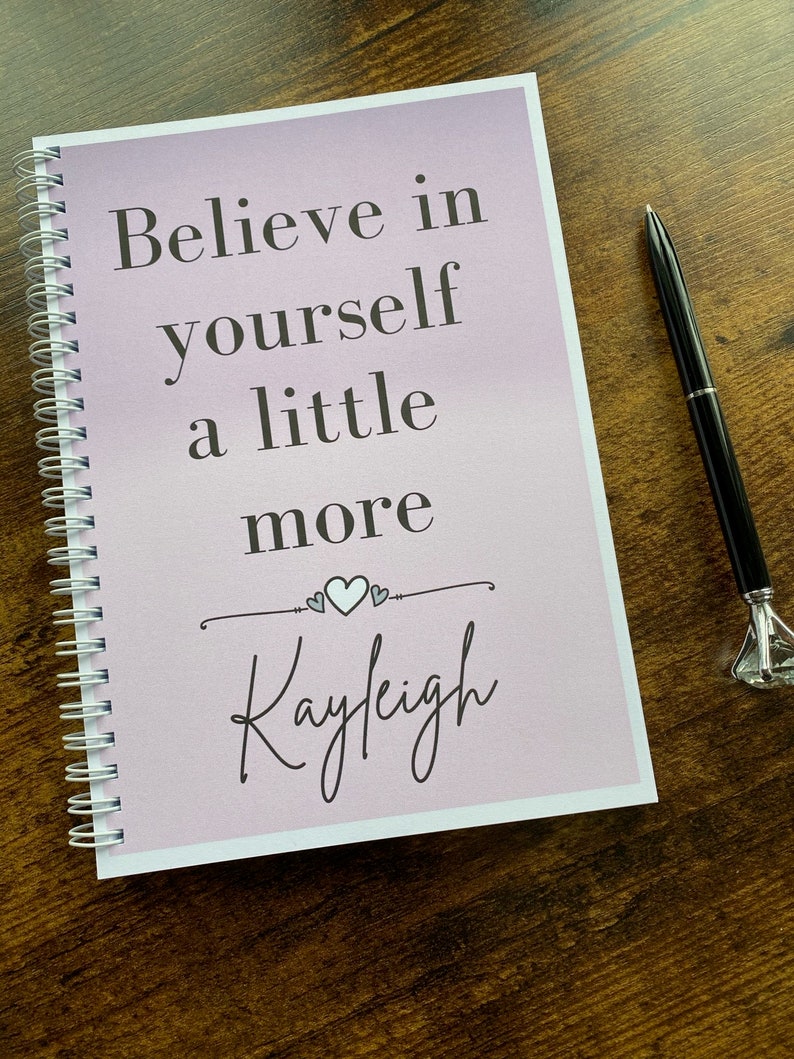 May include: A pink and white spiral bound journal with the text "Believe in yourself a little more" and the name "Kayleigh" written in cursive.