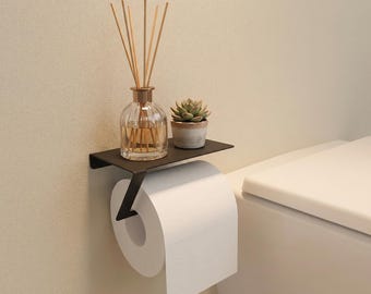 May include: A matte black metal toilet paper holder featuring a flat top shelf. A glass reed diffuser with wooden sticks and a small potted succulent sit on the shelf. A roll of white toilet paper hangs from the angled arm of the wall-mounted fixture.