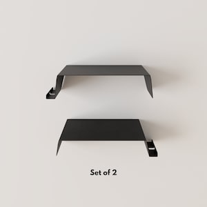Two black metal floating shelves with a minimalist design. Each shelf has a flat top and a downward-sloping front edge. The text "Set of 2" is displayed below the shelves.