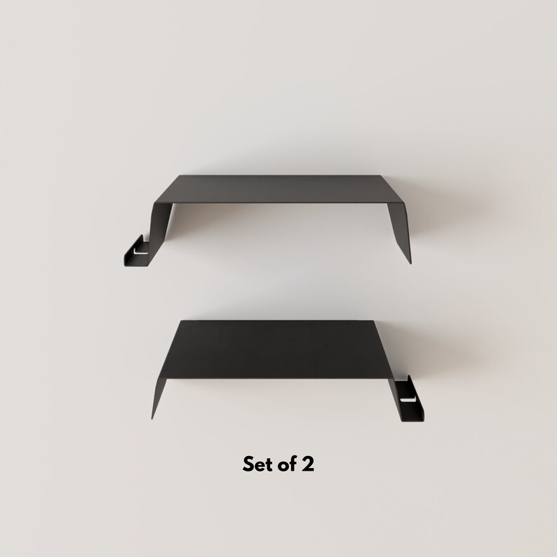 Two black metal floating shelves with a minimalist design. Each shelf has a flat top and a downward-sloping front edge. The text "Set of 2" is displayed below the shelves.