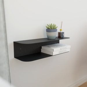 Könnte beinhalten: Ein schwarzes, schwebendes Regal aus Metall mit zwei Ebenen, an einer weißen Wand montiert. Die obere Ebene hat einen kleinen Topf mit einer Sukkulentenpflanze und eine Glasflasche mit einem Schilfrohrdiffusor. Die untere Ebene hat zwei Bücher übereinander gestapelt. Die Bücher sind mit "Arc Motions" und "Interior Design" betitelt.