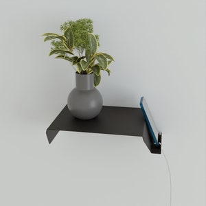 A black metal floating shelf mounted on a white wall, holding a gray vase with a plant. A phone is charging on the shelf. The shelf has a minimalist design.
