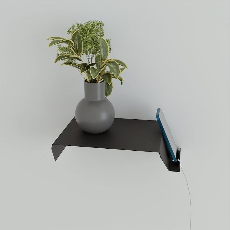 A black metal floating shelf mounted on a white wall, holding a gray vase with a plant. A phone is charging on the shelf. The shelf has a minimalist design.