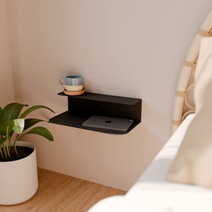 Könnte beinhalten: Ein schwebendes Regal aus schwarzem Metall mit zwei Ebenen, montiert an einer hellrosa Wand. Eine blaue und weiße Keramiktasse steht auf der oberen Ebene, und ein schwarzes Tablet-Computer befindet sich auf der unteren Ebene.