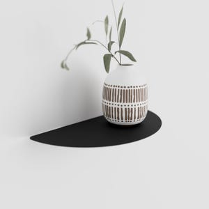 May include: A black half-circle shelf mounted on a white wall with a white vase with a brown geometric pattern holding a sprig of green leaves.