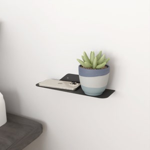 Puede incluir: Una estantería flotante de metal negro con borde redondeado, montada en una pared blanca. Un teléfono inteligente blanco y una pequeña planta suculenta en maceta con hojas verdes están en la estantería.