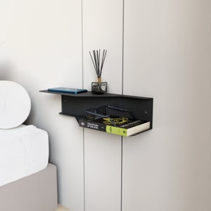 Puede incluir: Una estantería flotante de metal negro con un libro, gafas y un teléfono encima. La estantería está montada en una pared blanca.